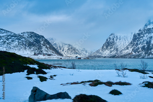 Remote winter coastal village nestled under majestic snow-covered mountains in Lofoten Islands, Northern Norway. Scenic Arctic fjord landscape during a Scandinavian road trip travel adventure.