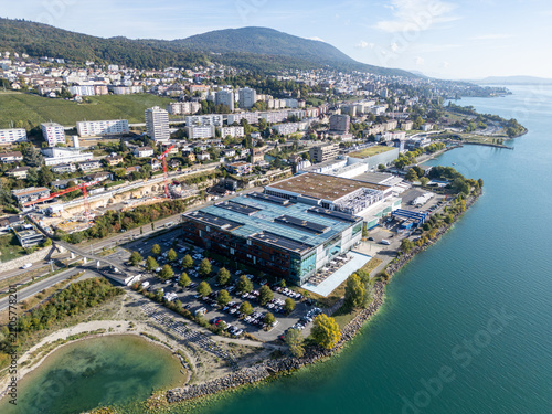 Aerial view of the Philip Morris RD Cube situated on the shore of Lake Neuchatel, surrounded by parking areas, urban development, and lush greenery in Neuchatel, Neuenburg, Switzerland.