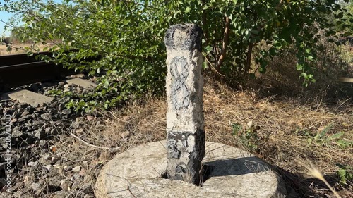 Weathered white and black concrete marker post by a railway track, camera pulls back to reveal dry grass, gravel and rails in sunlight