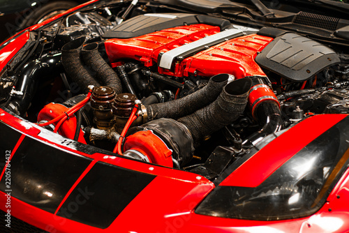 Red sports car engine with twin turbochargers, carbon fiber covers, black hoses, and glossy bodywork visible beneath an open hood in close up