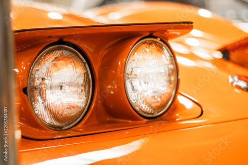 Close up of orange vintage car front with twin round headlights, glossy curved hood, chrome detailing, and reflections on classic automobile bodywork