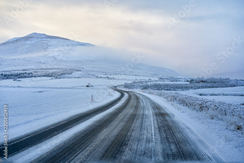 Scenic winding icy road curving through snow-covered landscape towards mountains in Iceland winter. Arctic highway road trip and self-drive travel adventure.
