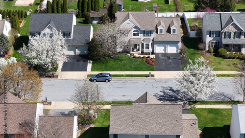 Aerial view of a suburban street featuring detached homes with manicured lawns, blooming spring trees, and a blue car on the road in Lititz, Pennsylvania, United States.