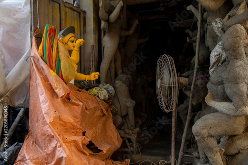 An artisan at work inside a traditional workshop in Kumartuli, Kolkata, preparing an unfinished, vibrant Durga idol during the monsoon season. The partially painted idol is wrapped in a plastic