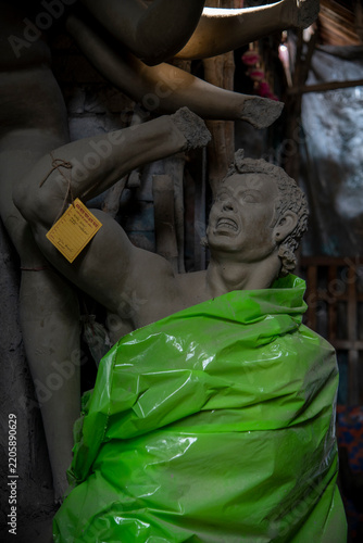 An unfinished, brightly colored Asur idol rests inside a traditional artisan workshop in Kumartuli, Kolkata. Partially covered with a clear plastic sheet to protect the intricate clay