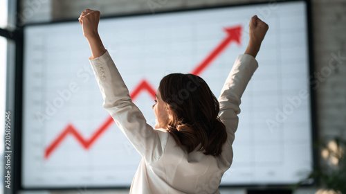 Businesswoman celebrating with raised arms against rising red trend arrow graph, financial success and stock market growth achievement concept image