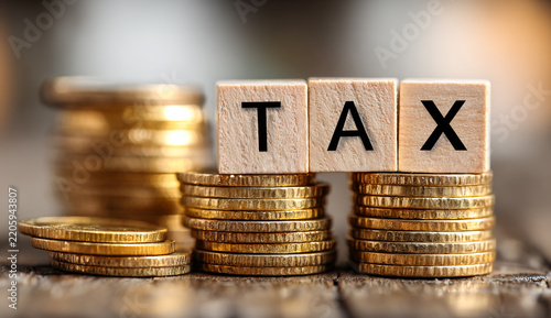 Wooden blocks spelling the word TAX resting on stacks of golden coins, representing financial planning and taxation concept.