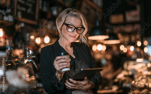 Smiling shop owner woman wearing glasses and apron holding cup in cafe with warm bokeh background, friendly hospitality and small business concept portrait