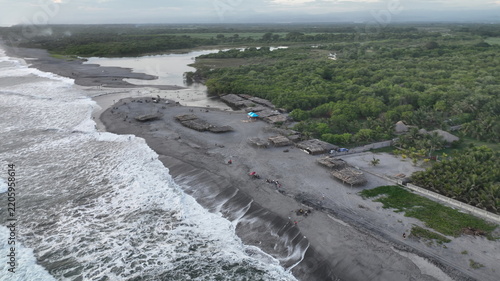 Aerial view of San Marcelino beach with dark volcanic sand, crashing Pacific waves, a coastal lagoon, and thatched-roof huts amidst lush vegetation in La Paz Centro, La Paz, El Salvador.