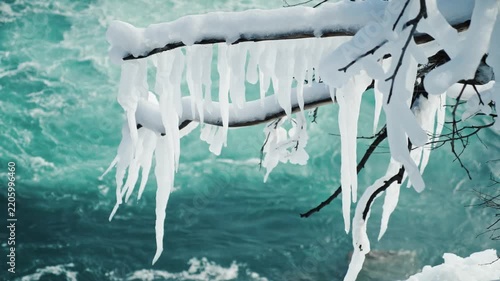 Close up view of snow-covered branches and icicles dangling above a vibrant blue river, dynamic winter scenery with flowing water.
