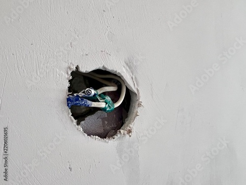 Circular drywall cutout with exposed electrical wires wrapped in tape on a light wall, copy space for text.