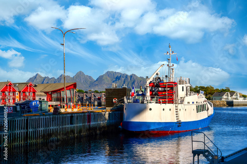 Ship moored in Svolvaer harbor in the Lofoten Islands, with colorful houses and distinctive mountain scenery beneath a clear blue sky.