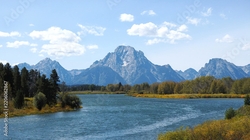Serene river flows past towering mountain peaks. Grand Tetons, Oxbow Bend. Golden reeds line the water's edge. Evergreen trees frame the scene below. Clouds drift softly across the sky