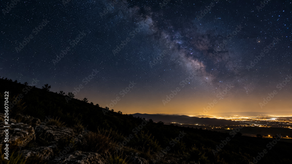 Fototapeta premium Milky Way galaxy over night landscape with distant glowing city lights on horizon. Concept of starry sky, astrophotography, stars, night sky. Copy space.