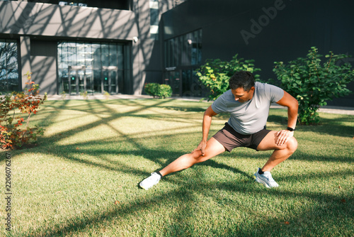 Adult man performing a side lunge stretch on a sunny lawn near a modern building. Outdoor fitness and flexibility exercise with copy space