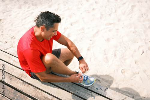 Athletic man ties running shoes while sitting on a wooden bench beside sand, preparing for an outdoor workout, cardio training or run in warm sunny weather.