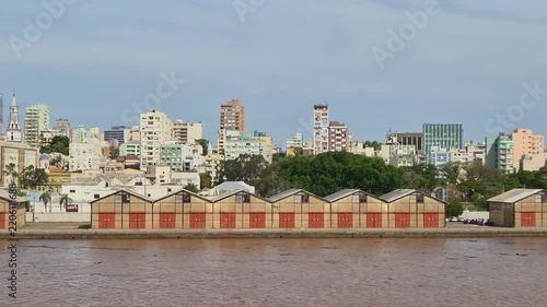 Video shot panning across waterfront port warehouses along a wide brown river, backed by a dense cityscape of high-rise buildings and green trees under a bright blue afternoon sky