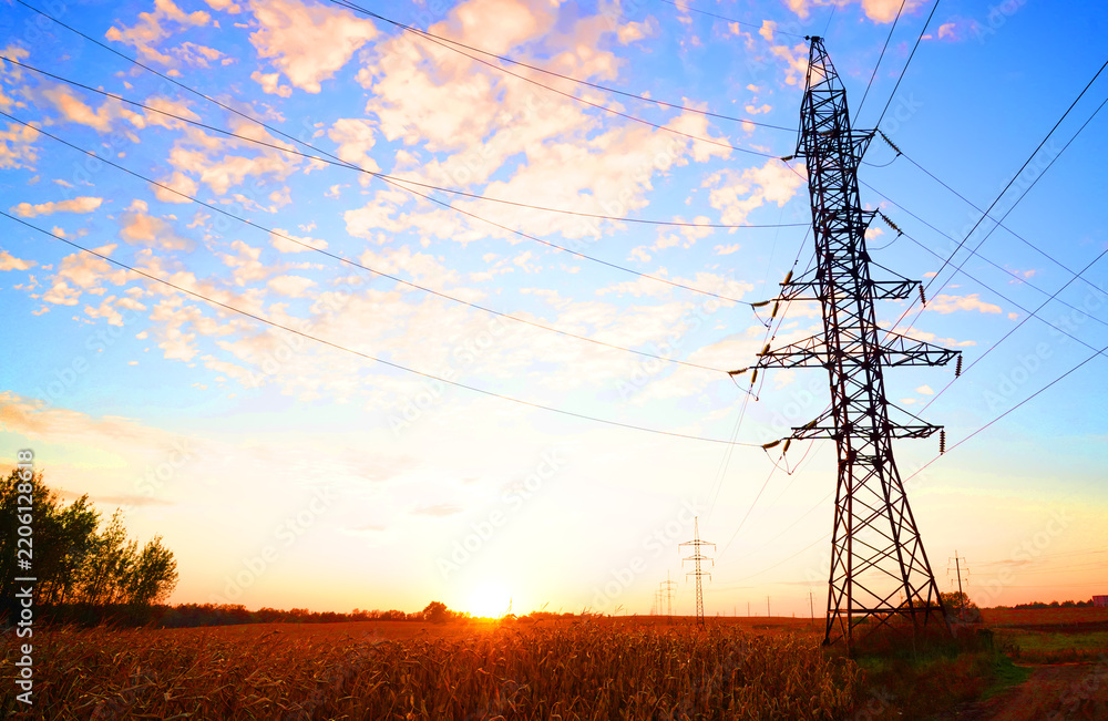 Obraz premium Silhouetted electricity pylon in golden field at sunset with blue sky and scattered clouds, power lines stretching across horizon