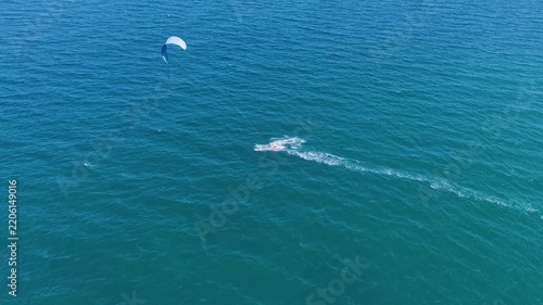 Dynamic kitesurfing action in the ocean. Aerial view of many people riding the waves on the weekend. Colorful parachutes of surfers against the blue sea and sky. Kiteboarding.