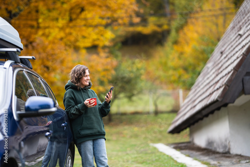 A smiling woman standing next to a blue car with a roof box, holding a red mug and using a smartphone in front of an A-frame house in autumn