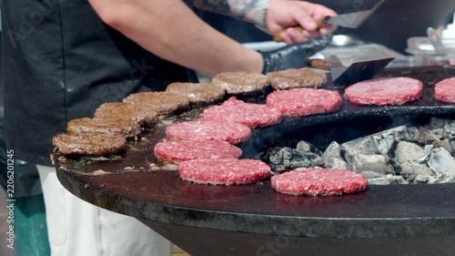 Cook turns burger patties on hot grill. Smoke rises from burning charcoal beneath. Fresh meat sizzles as it cooks evenly. Grill has many patties, some brown, some raw