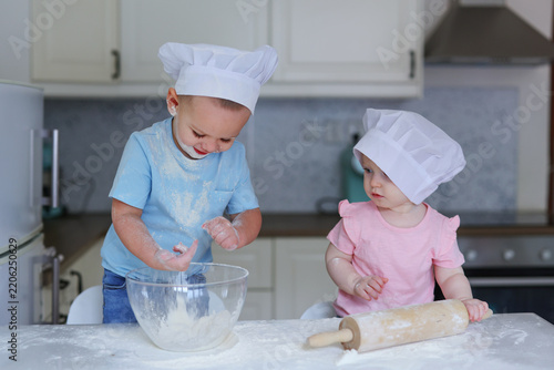 Two children in the kitchen pour out the flour and dressed in the form of cooks. Children's games in adult professions. Photo in interior.