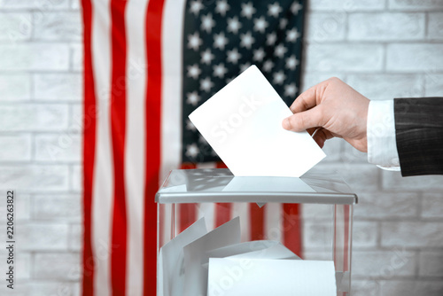 Voter casting a voting ballot into a transparent ballot box with the US flag. American elections, democracy and voting rights.