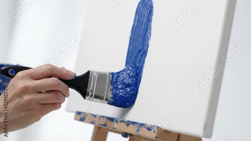 Hand applying blue paint on canvas with paintbrush on easel in bright studio