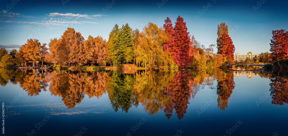 Obraz premium Panoramic autumn view of Boating Island in Szombathely town, administrative centre of Vas County, Hungary. Calm afternoon scene of Boating lajke. Trees covered with orange and crimson leaves.