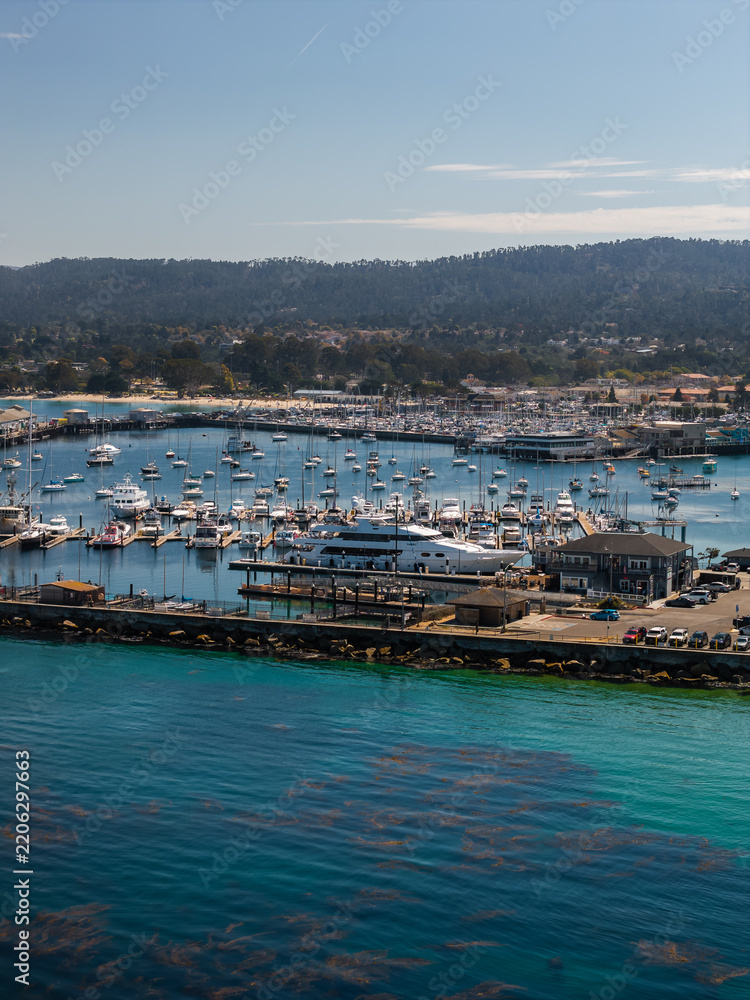 Fototapeta premium Aerial view shows boats moored along docks at Monterey Harbor in California, with the coastal shoreline, breakwater, and surrounding forested hills visible under daylight.