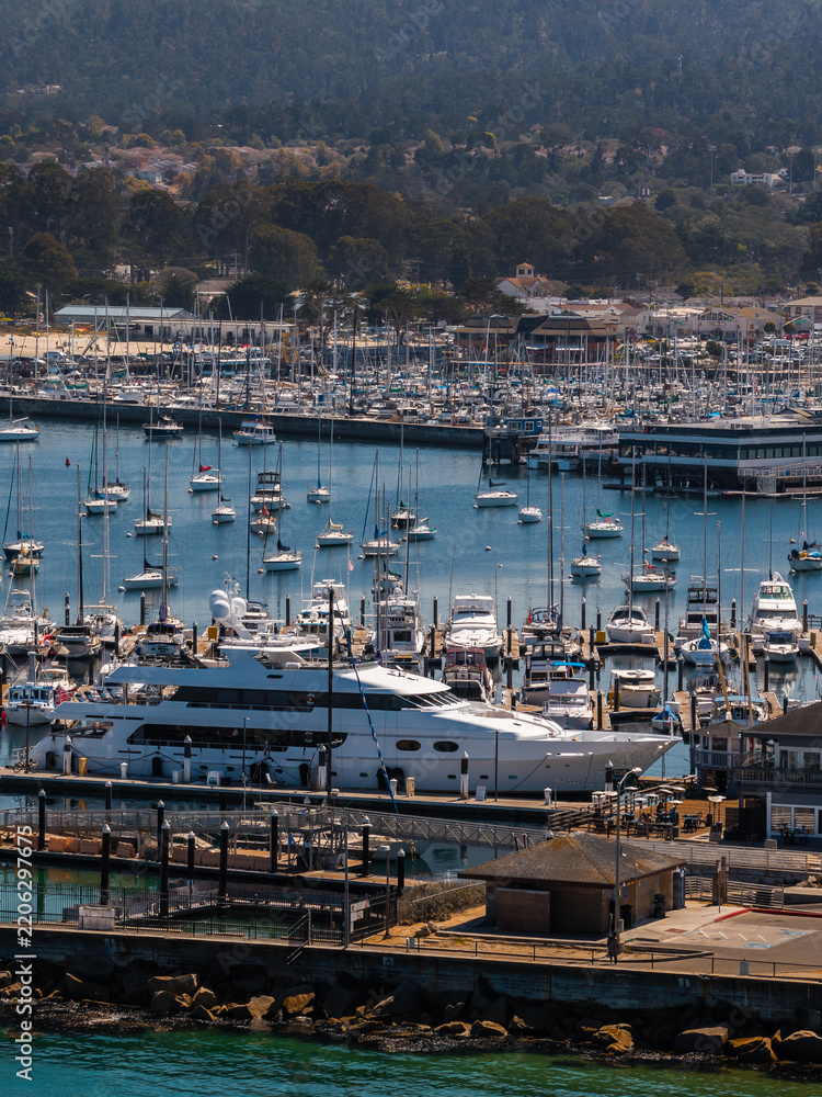 Fototapeta premium Aerial shot displays numerous sailboats, yachts, and leisure watercraft moored along the docks and marina in Monterey, California, with coastal buildings visible inland.