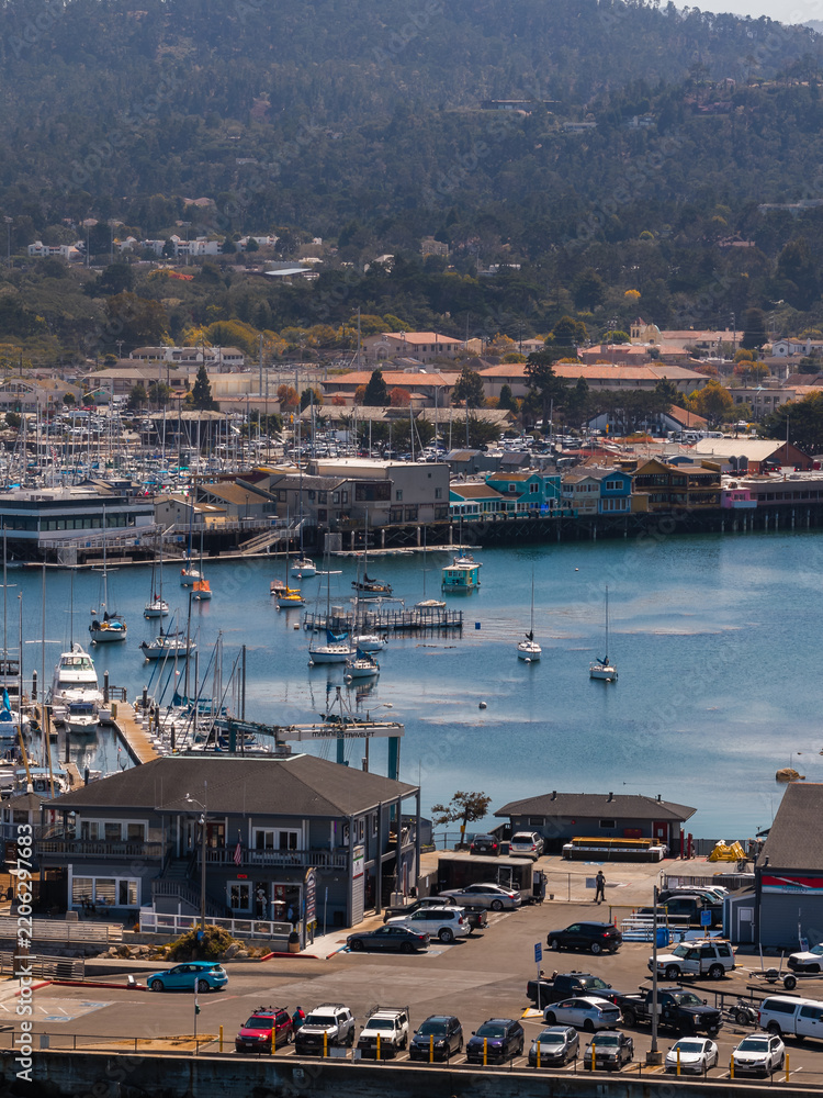 Fototapeta premium Aerial shot of Monterey harbor displaying moored sailboats, Fisherman's Wharf buildings, a parking lot, and forested hills under natural daylight.