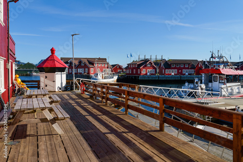 Sunny harbor scene in Svolvaer on the Lofoten Islands with a wooden terrace, boats and traditional red wooden houses beside the water under clear blue sky.