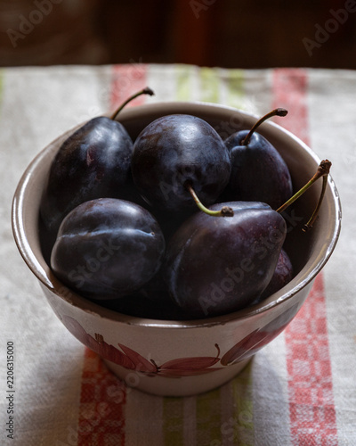Fresh dark plums in a rustic hand-painted ceramic bowl on a striped linen towel