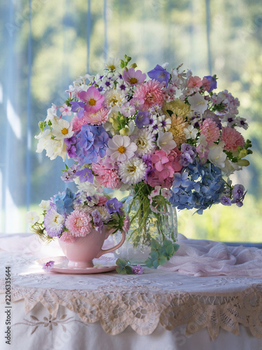 Beautiful bouquet in a vase flowers phlox, asters, hydrangeas and bellflowers on a table, cup and saucer, still life set against a window overlooking the garden.
