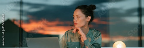 Woman working at laptop looking thoughtful during sunset through a window for remote work background and contemplative business banner