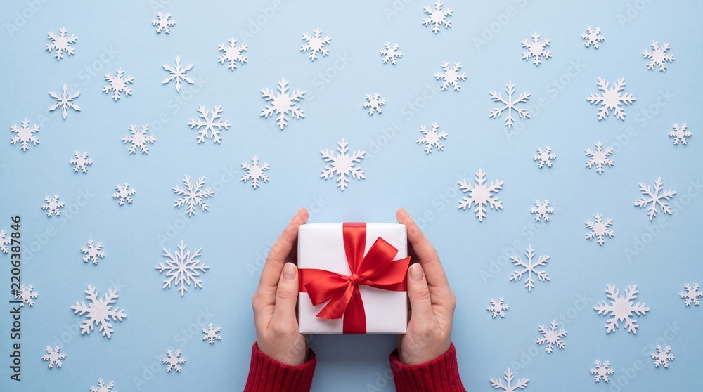 Obraz premium High angle view of human hands in red sweater holding a white gift box with a red ribbon on a light blue background decorated with white snowflakes, winter holiday celebration and giving concept.