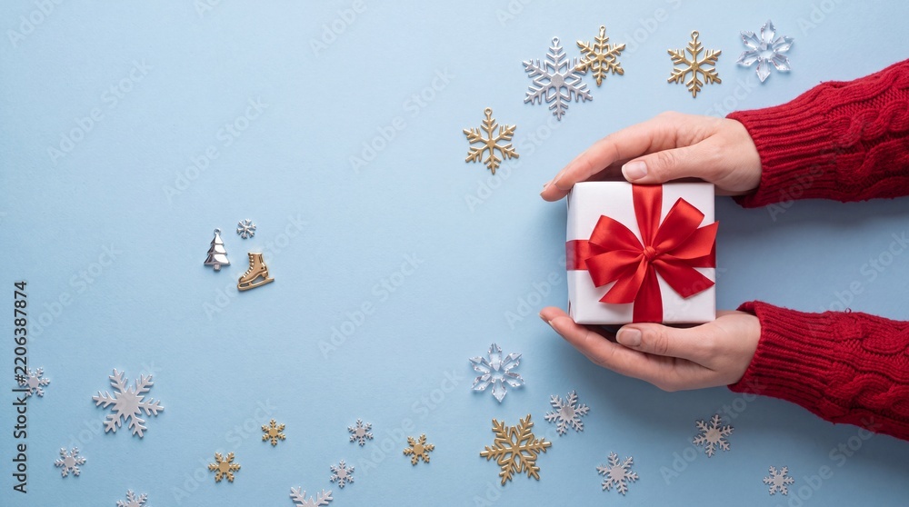 Obraz premium Top View of Woman Hands in Red Knitted Sweater Holding a White Christmas Gift Box with Red Ribbon Bow Surrounded by Snowflake Decorations and Winter Charms on Pastel Blue Background with Copy Space