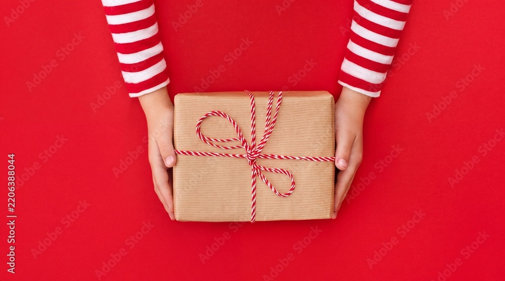 Obraz premium Top view of a child's hands wearing festive red and white striped sleeves holding a handmade gift box wrapped in brown kraft paper and tied with red twine over a vibrant red background layout.