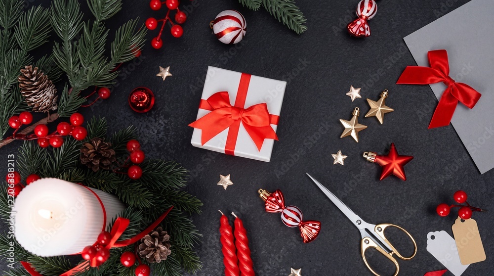 Obraz premium Top view of festive Christmas holiday preparation featuring a white gift box with red ribbon, burning candle, pine branches, red berries, and golden ornaments on dark background for seasonal DIY.