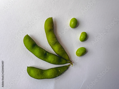 Fresh green edamame pods rest on plain white surface. Three pods curve gently, still attached to stem. Three peas lie scattered nearby, vibrant and plump