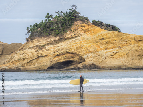 A surfer carrying a yellow longboard walks along the wet sand at Pacific City Beach beneath dramatic golden sandstone cliffs on the Oregon Coast