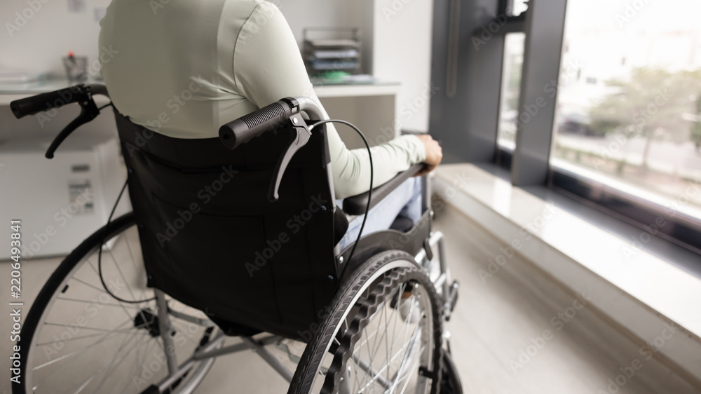 Fototapeta premium Close up cropped image rearview, unrecognizable young woman with disability sitting in wheelchair looking out window in medical office, getting accessible healthcare services in rehabilitation center