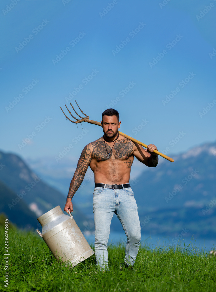 Obraz premium Farmer carrying milk can on grassy hillside. Handsome farmer with pitchfork beside mountain lake. Farmer with bare chest in rural portrait. Tattooed dairy farmer carrying farm tool outdoors.