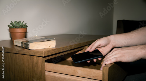 A pair of hands places a smartphone face-down into a wooden drawer with a book and plant visible nearby in soft natural light