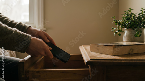 A person places their smartphone face-down into a wooden drawer with a book and plant nearby under calm natural lighting