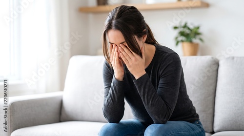 A young woman sits on a couch, covering her face with her hands, appearing distressed or sad.