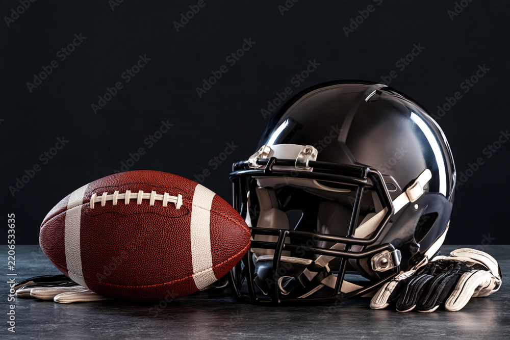 Obraz premium Black helmet rests beside a textured football. Gloves lie nearby, reflecting team spirit. Dark background highlights gear's bold design. Image conveys athletic readiness and focus