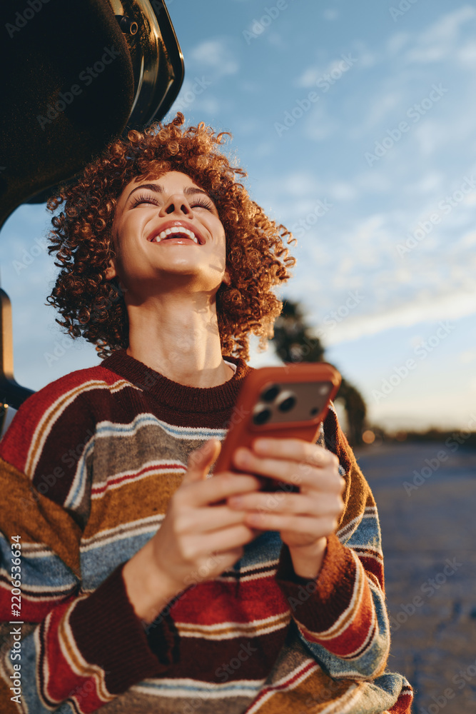 Obraz premium Woman with curly hair smiling outdoors, holding smartphone near road at sunset, wearing striped sweater, golden evening light, casual lifestyle, candid street portrait, relaxed mood