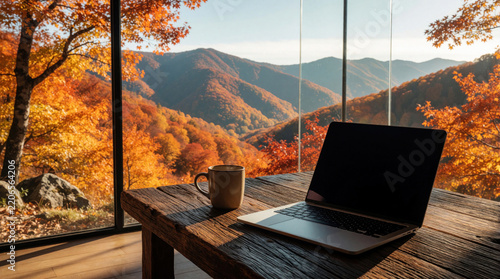 Laptop, coffee mug sit on rustic wooden table near large window. Autumn mountain landscape with colorful orange trees views from cozy cabin. Digital nomad works remote in peaceful forest retreat
